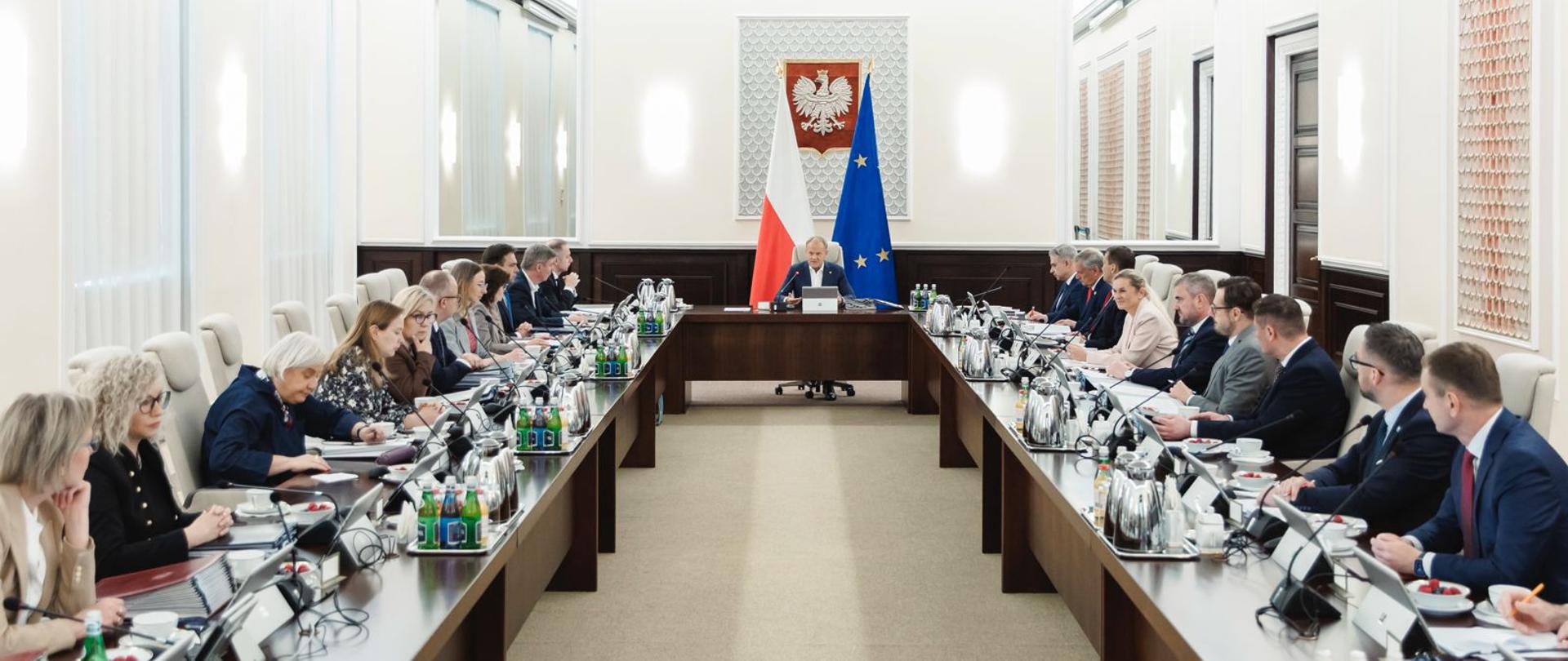 Obraz przedstawia długi stół konferencyjny w kształcie litery „U” w dużej, formalnej sali. Wokół stołu siedzą liczni dorośli, prawdopodobnie na spotkaniu. Na ścianie za centralnym miejscem przy stole wiszą polskie i europejskie flagi oraz godło Polski. Na stole znajdują się dokumenty, laptopy, butelki z wodą i szklanki. Pomieszczenie ma białe ściany i eleganckie, klasyczne wykończenia.