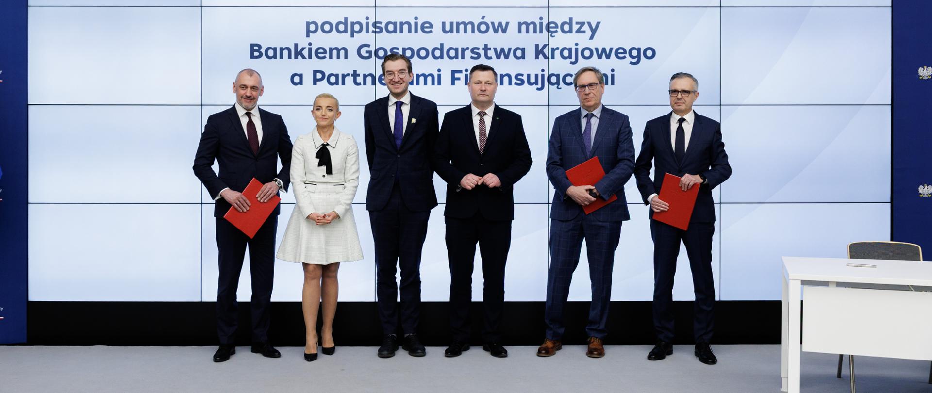 W sali konferencyjnej sześć osób stoi na scenie i pozuje do zdjęcia. Trzeci od lewej wiceminister Jan Szyszko.