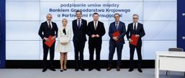 W sali konferencyjnej sześć osób stoi na scenie i pozuje do zdjęcia. Trzeci od lewej wiceminister Jan Szyszko.