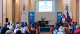 Szerokie ujęcie sali konferencyjnej, w której odbywa się Doroczna Konferencja Sieci Szkół Stowarzyszonych UNESCO w Polsce. Uczestnicy siedzą na czerwonych krzesłach. Na środku stoi mównica z mówcą i ekran, na którym wyświetlana jest prezentacja UNESCO. Widać flagi i rollupy UNESCO oraz ORE.