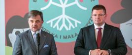 Z przodu sali stoi mężczyzna w garniturze, mówi do mikrofonu na stojaku, obok niego wiceminister Rzymkowski, za nimi ekran z napisem Bez korzeni nie zakwitniesz. Moja Wielka i Mała Ojczyzna.