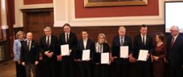 10 osób stoi przed stołem, pokazują trzymane w rękach dokumenty, za nimi na czerwonej ścianie obraz.