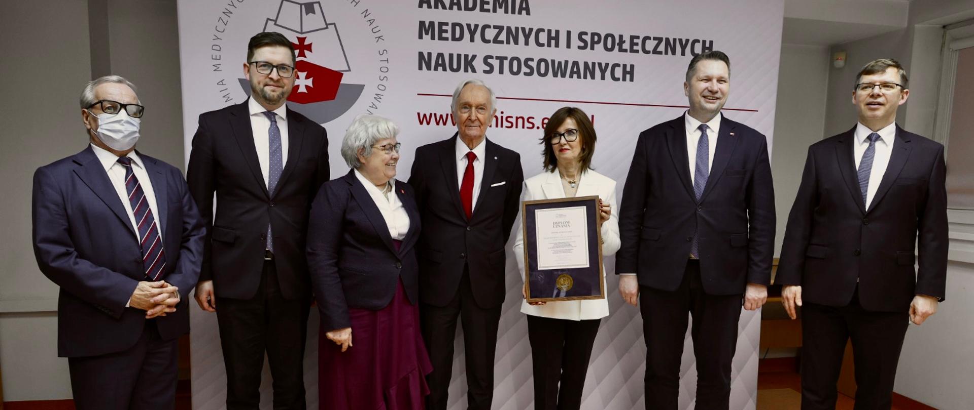 Pod białą ścianką z napisem Akademia medycznych i społecznych nauk stosowanych stoi siedem uśmiechniętych osób, kobieta w białej marynarce pokazuje dokument w ramkach.