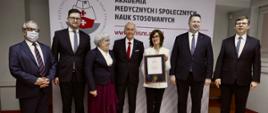 Pod białą ścianką z napisem Akademia medycznych i społecznych nauk stosowanych stoi siedem uśmiechniętych osób, kobieta w białej marynarce pokazuje dokument w ramkach.