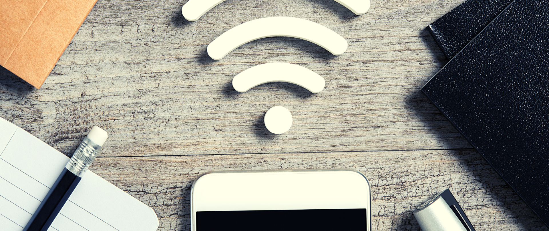 Biurko. Na środku widać gorą część białego smartfona a nad nim symbol siedzi wi-fi. Po lewej stronie leżą czarne teczki, po prawej u góry notesik z brązową okładką, na dole otwarty notes w linie a na nim ołówek.