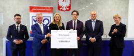 Pod szarą ścianą przed niebieskim stołem stoi sześć osób, kobieta i mężczyzna pośrodku trzymają wielki symboliczny czek z napisem Gmina Kozienice - 1 000 000 zł.