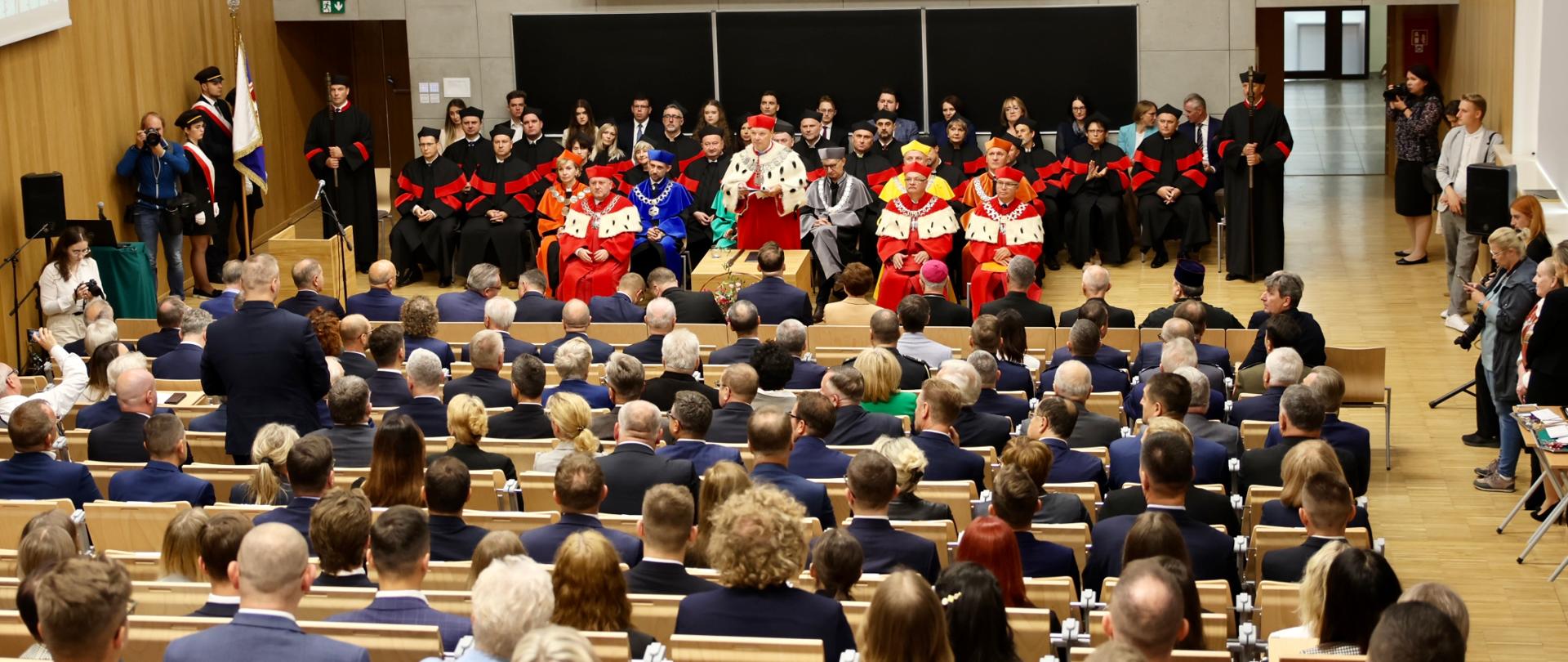 Widok z góry na dużą aulę, w ławach siedzą ludzie, przed nimi z przodu sali w kilku rzędach krzeseł siedzą ludzie w ceremonialnych czerwonych i czarnych strojach.