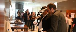 Pod ścianą stoją stoliki, na nich laptopy, przy stolikach stoją ludzie w okularach VR.