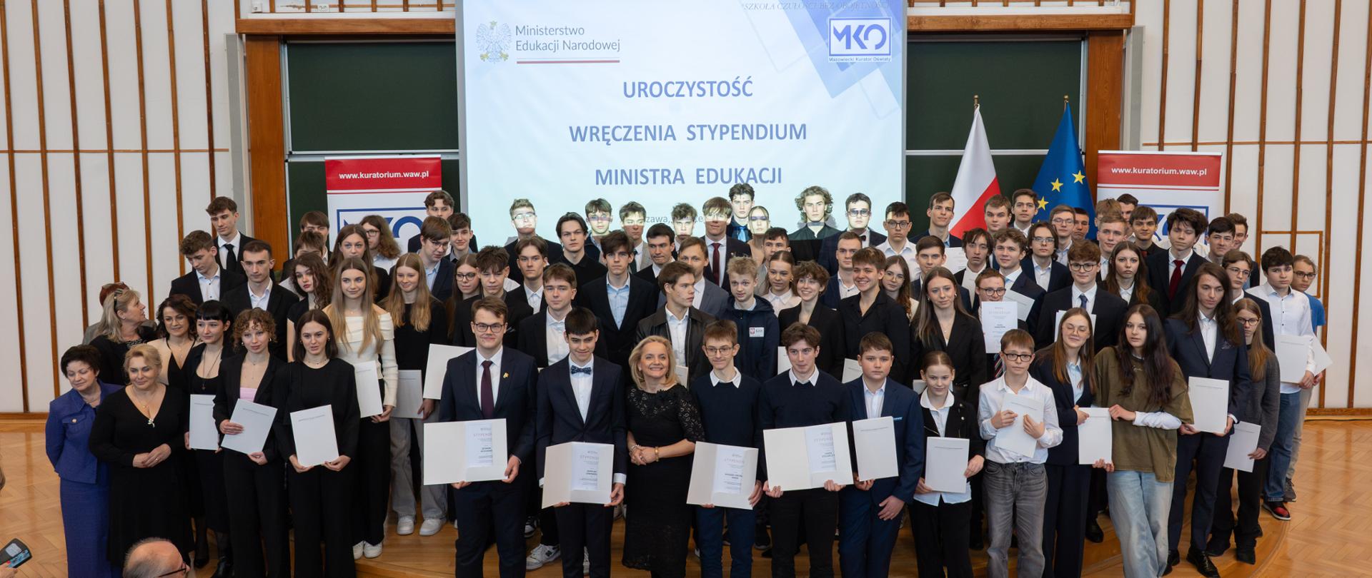 Zdjęcie zbiorowe . Na scenie stoi duża grupa uczniów z otwartymi dokumentami i pozuje do zdjęcia. W tle ekran z napisem Uroczystość wręczenia stypendiów Ministra Edukacji
