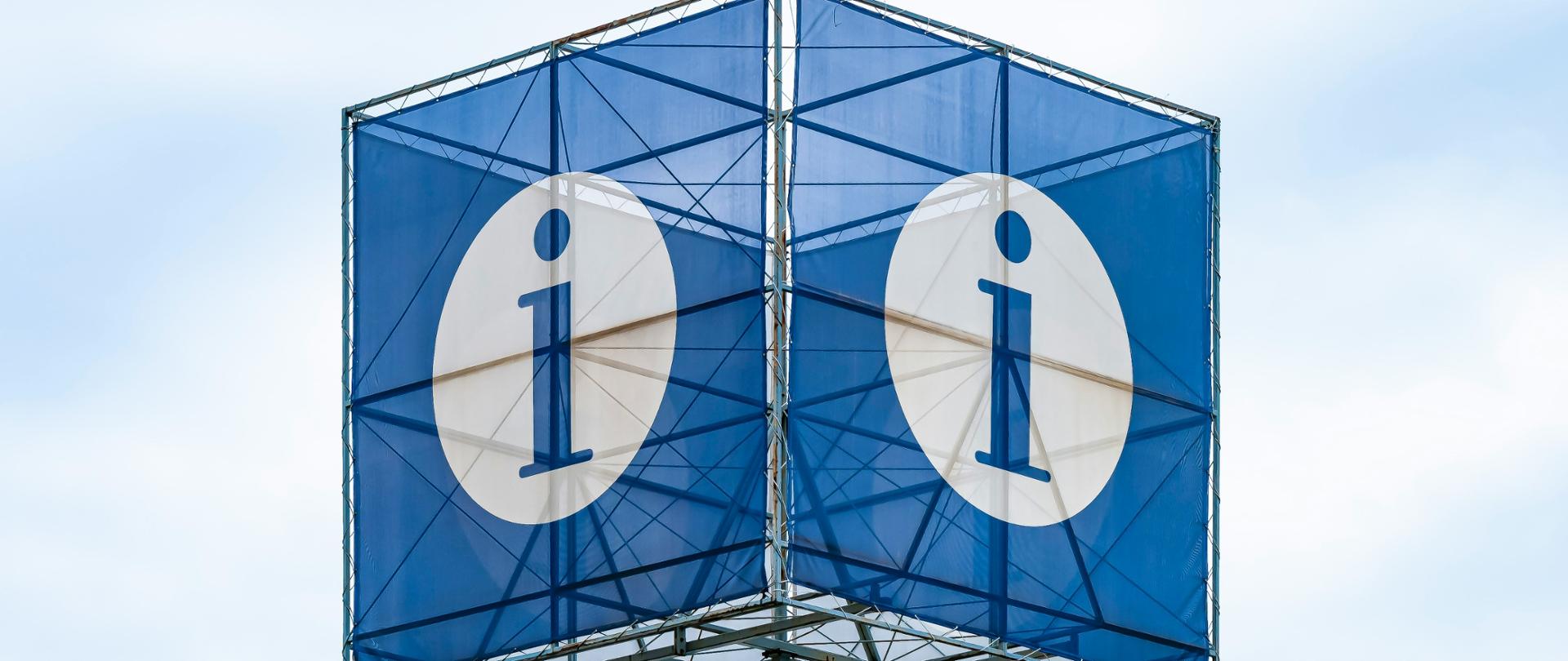 Na metalowym słupie wiszą dwa niebieskie banery na środku biała kropka z niebieską literą i symbolizującą informację