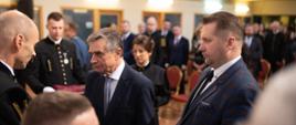 Minister Czarnek i stojący obok starszy mężczyzna w garniturze rozmawiają z ludźmi w czarnych galowych mundurach, minister trzyma w ręku otwartą małą książeczkę, dalej sala z żółtymi ścianami wypełniona ludźmi.