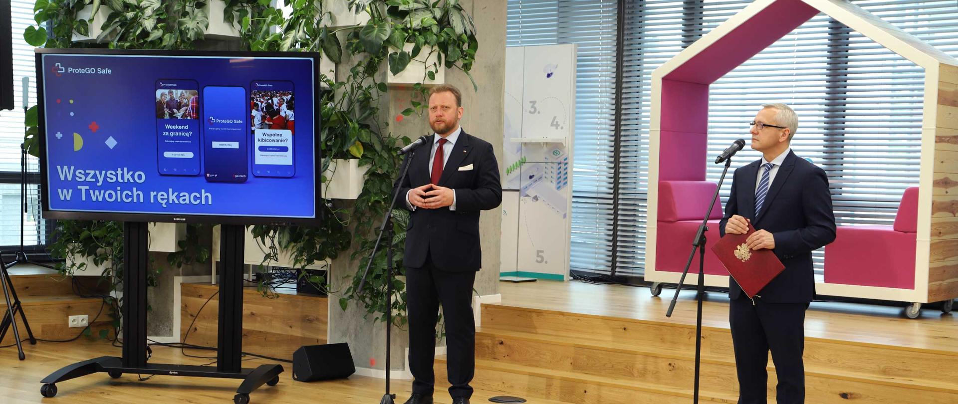 Zdjęcie podczas konferencji poświęconej aplikacji ProteGo Safe. Po prawej stronie Minister Cyfryzacji - Marek Zagórski przy mikrofonie, w ręku trzyma teczkę z godłem Polski. Po lewej stronie Minister Zdrowia - Łukasz Szumowski przed mikrofonem. Obok ministra Szumowskiego na ekranie telewizora wyświetlona grafika z napisem "Wszystko w Twoich rękach" 