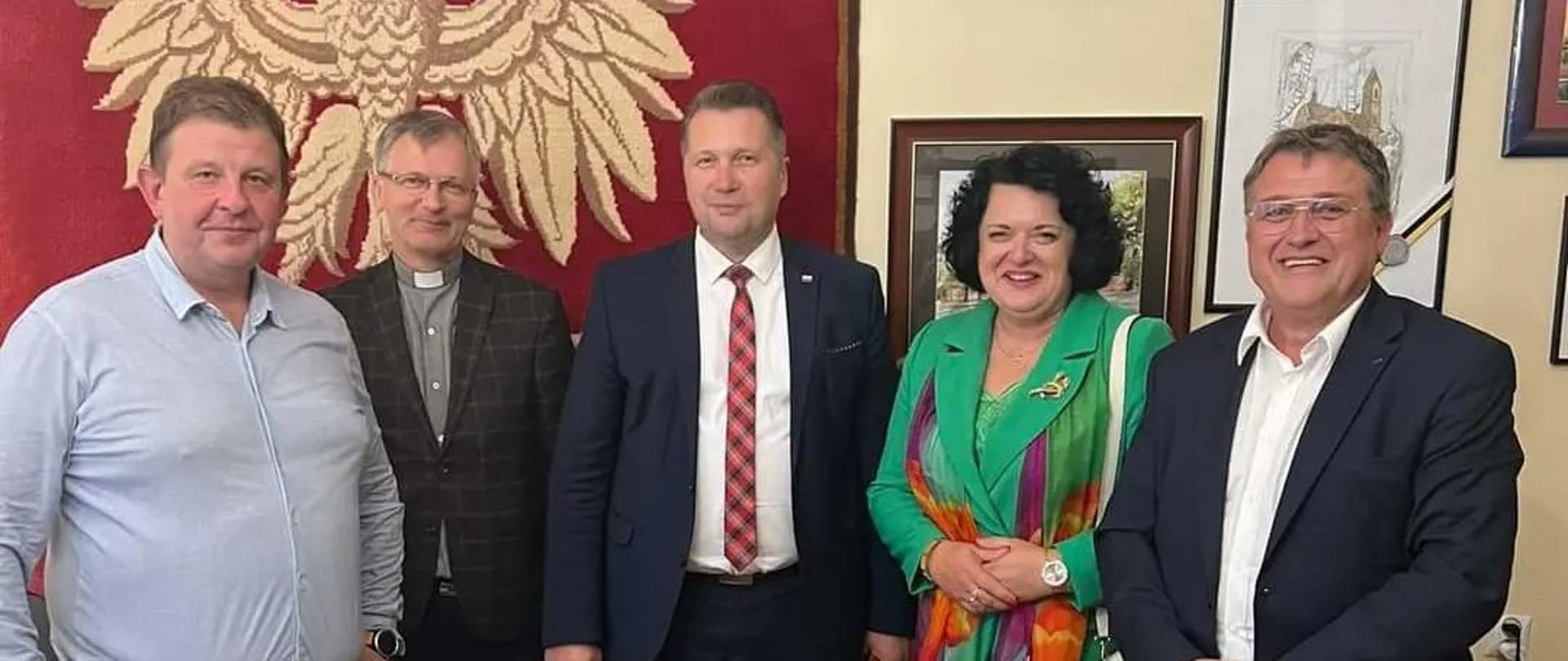 Minister w otoczeniu kilku osób stoi pod ścianą, na której wisi wielkie godło Polski.