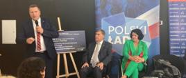 Minister stoi i mówi do mikrofonu, za nim tablica z napisem Polski ład, obok i przed nim siedzą ludzie na krzesłach.