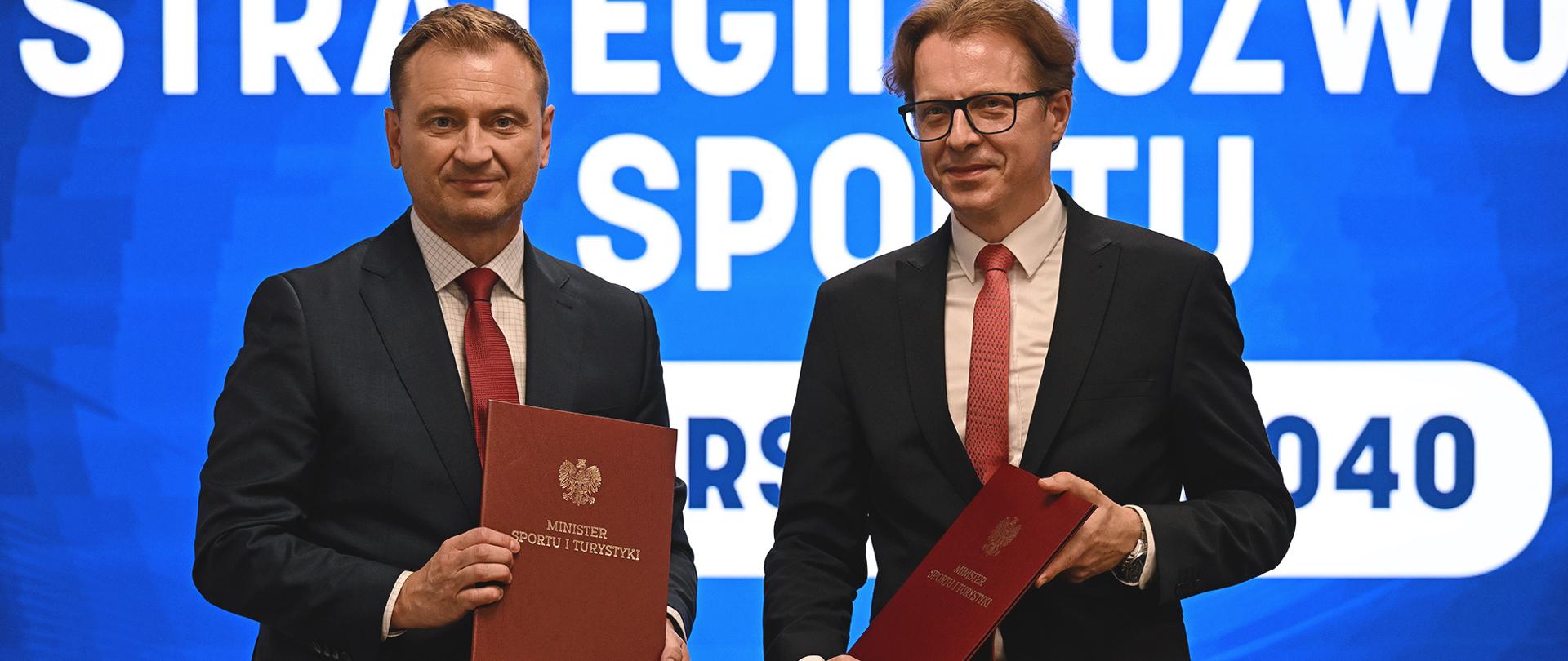 Na zdjęciu od lewej: Minister Sportu i Turystyki Sławomir Nitras i Dionizy Smoleń Partner w PwC Polska po podpisaniu umów na realizację "Strategii Rozwoju Sportu - IO Warszawa 2040". W tle ekran z wyświetloną grafiką z niebieskim tłem.