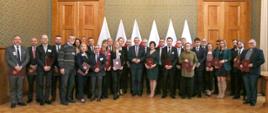 Zdjęcie zbiorowe, na drewnianej podłodze tle zielonej ściany wyłożonej drewnianymi panelami stoi duża grupa elegancko ubranych osób, trzymają w rękach czerwone teczki ze złotym godłem.