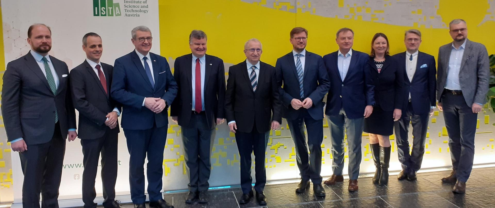 Na podłodze z szarych kafli, przed żółtą ścianą w szary wzorek stoi w rzędzie 10 elegancko ubranych osób.