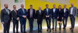 Na podłodze z szarych kafli, przed żółtą ścianą w szary wzorek stoi w rzędzie 10 elegancko ubranych osób.
