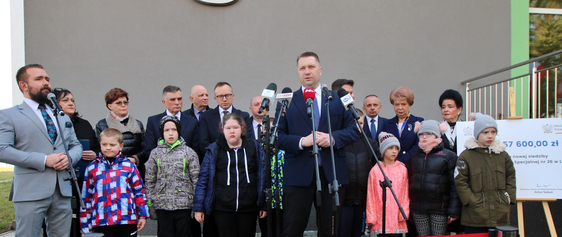 Na wyłożonym szarą kostką podwórku stoi minister Czarnek mi mężczyzna w jasnoszarym garniturze, obaj mają przed sobą mikrofony na stojakach, za nimi szereg kolorowo ubranych dzieci, dalej pod szarą ścianą grupa dorosłych, z przodu między dwoma metalowymi słupkami czerwona taśma.