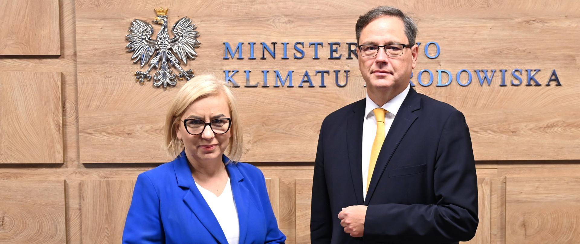 Dwie osoby stoją przed ścianą z godłem Polski i napisem „Ministerstwo Klimatu i Środowiska”. Osoba po lewej ma niebieską marynarkę i okulary, osoba po prawej ciemny garnitur, żółty krawat i okulary.