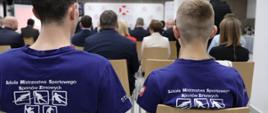 Uczniowie Szkoły Mistrzostwa Sportowego Rekord, dwóch chłopców siedzi tyłem, ubrani są w niebieskie koszulki z napisem szkoła Mistrzostwa Sportowego Sportów Zimowych