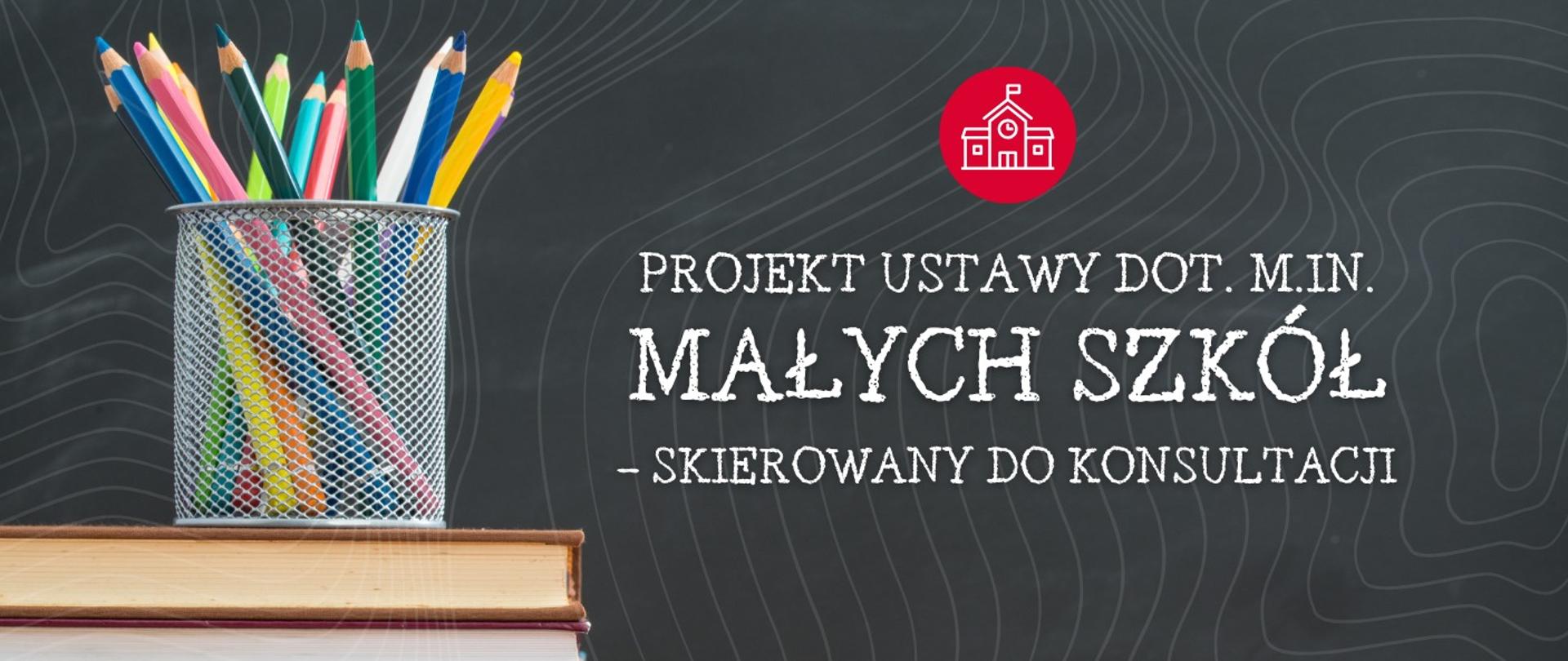 Przedstawiono biurko szkolne z otwartym pojemnikiem na ołówki w różnych kolorach i stos książek. Tło stylizowane jest na tablicę szkolną. W prawym górnym rogu znajduje się ikona szkoły w czerwonym kółku. Obok ikony jest napis „PROJEKT USTAWY DOT. M.IN. MAŁYCH SZKÓŁ - SKIEROWANY DO KONSULTACJI”.