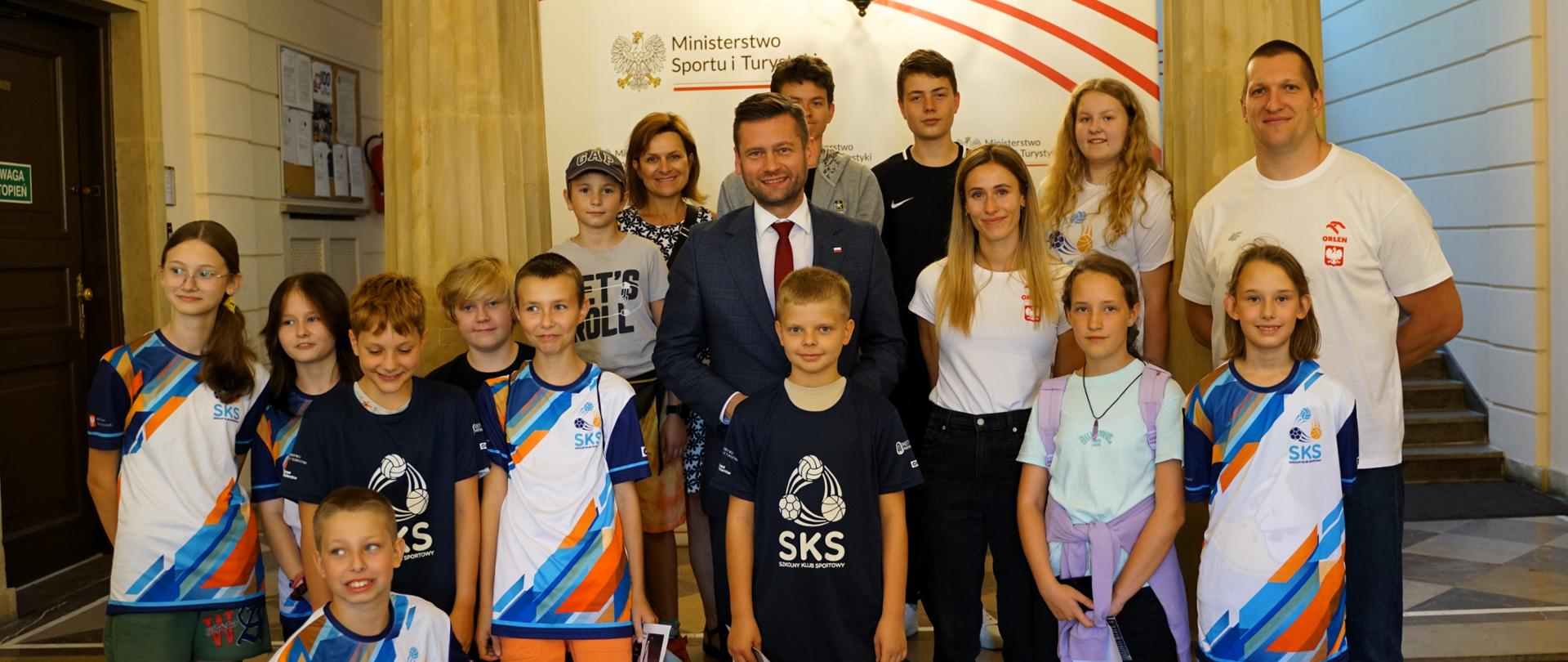 Na środku zdjęcia stoi minister Kamil Bortniczuk z Natalią Kaczmarek i Wojciechem Nowickim. Wokół ministra i sportowców stoją dzieci ubrane w koszulki SKS, szkolny klub sportowy. W widać białą ściankę z napisem Ministerstwo Sportu i Turystyki oraz orłem w kornie. 