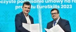 Dwóch mężczyzn w garniturach stoi na tle niebieskiej ściany, każdy trzymaj w ręku otwartą teczkę z dokumentami, podają sobie dłonie.