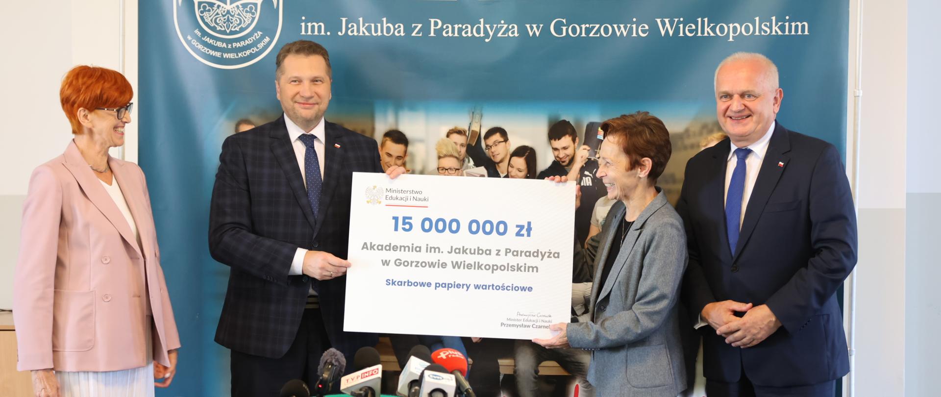 Dwie kobiety i i dwóch mężczyzn stoją na tle baneru z napisem Akademia im. Jakuba z Paradyża w Gorzowie Wielkopolskim. Minister Czarnek wraz z jedną z kobiet trzyma symboliczny czek z napisem: 15 mln zł Akademia im. Jakuba z Paradyża w Gorzowie Wielkopolskim. 
