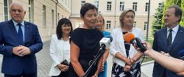 Na dworze, przed starym budynkiem stoi grupa ludzi, z przodu kobieta w granatowej sukience mówi do dwóch mikrofonów trzymanych przez reporterów.
