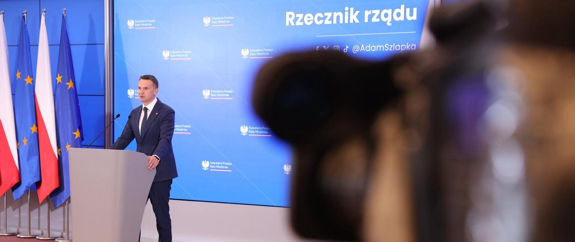Po lewej stronie zdjęcia stoi Rzecznik rządu Adam Szłapka podczas konferencji prasowej po posiedzeniu Rady Ministrów, za szarą mównicą. Za nim widoczny jest niebieski ekran, wyświetlający logo KPRM, na lewo od rzecznika widoczne są flagi Polski i Unii Europejskiej. W prawej części zdjęcia widoczna jest rozmazana kamera telewizyjna, nagrywająca rzecznika.