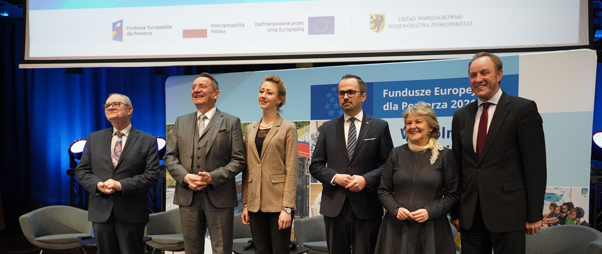 Sześć osób stoi na scenie i się uśmiecha