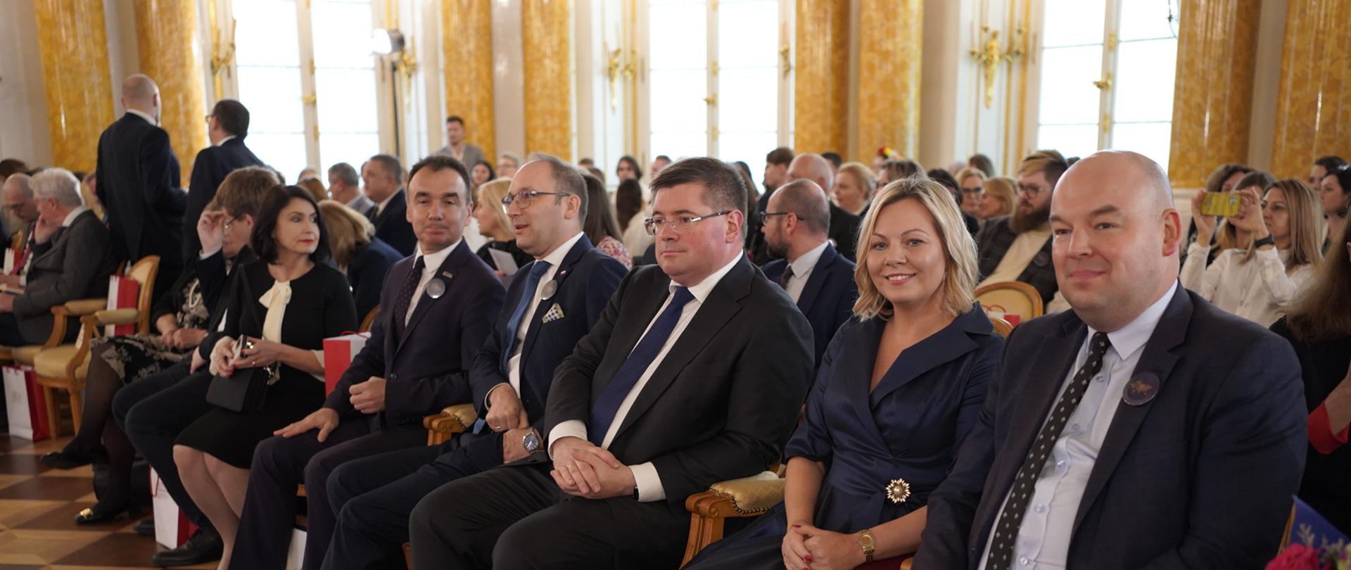 Widok z boku na wypełnioną siedzącymi na krzesłach ludźmi salę, w pierwszym rzędzie wiceminister Rzymkowski, w tle ściany zdobione złoconymi kolumnami.