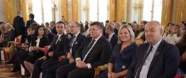 Widok z boku na wypełnioną siedzącymi na krzesłach ludźmi salę, w pierwszym rzędzie wiceminister Rzymkowski, w tle ściany zdobione złoconymi kolumnami.