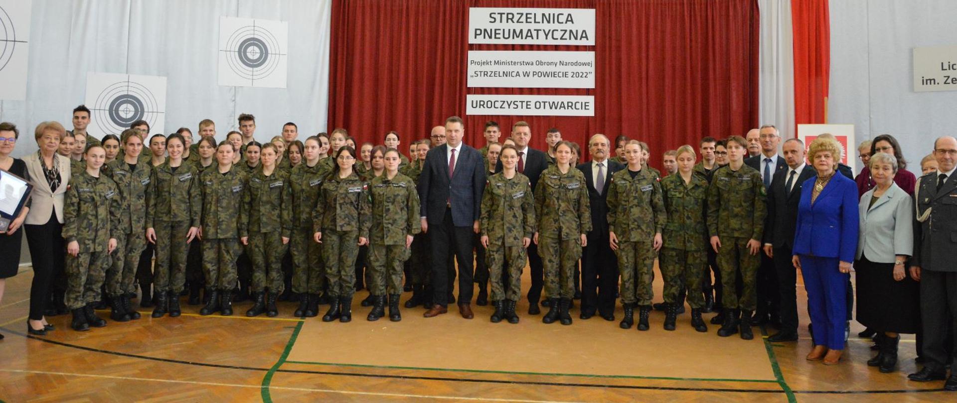 W dużej sali, pod ścianą przykrytą czerwoną kotarą z napisem Strzelnica pneumatyczna stoi duża grupa młodzieży w polskich mundurach w kamuflażu wz. 93, pośrodku grupy minister Czarnek.