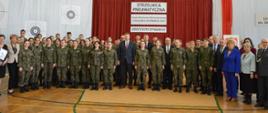 W dużej sali, pod ścianą przykrytą czerwoną kotarą z napisem Strzelnica pneumatyczna stoi duża grupa młodzieży w polskich mundurach w kamuflażu wz. 93, pośrodku grupy minister Czarnek.