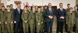 Na sali z drewnianą podłogą stoi duża grupa młodzieży w zielonych mundurach, między nimi wiceminister Murdzek i dwóch mężczyzn w garniturach.