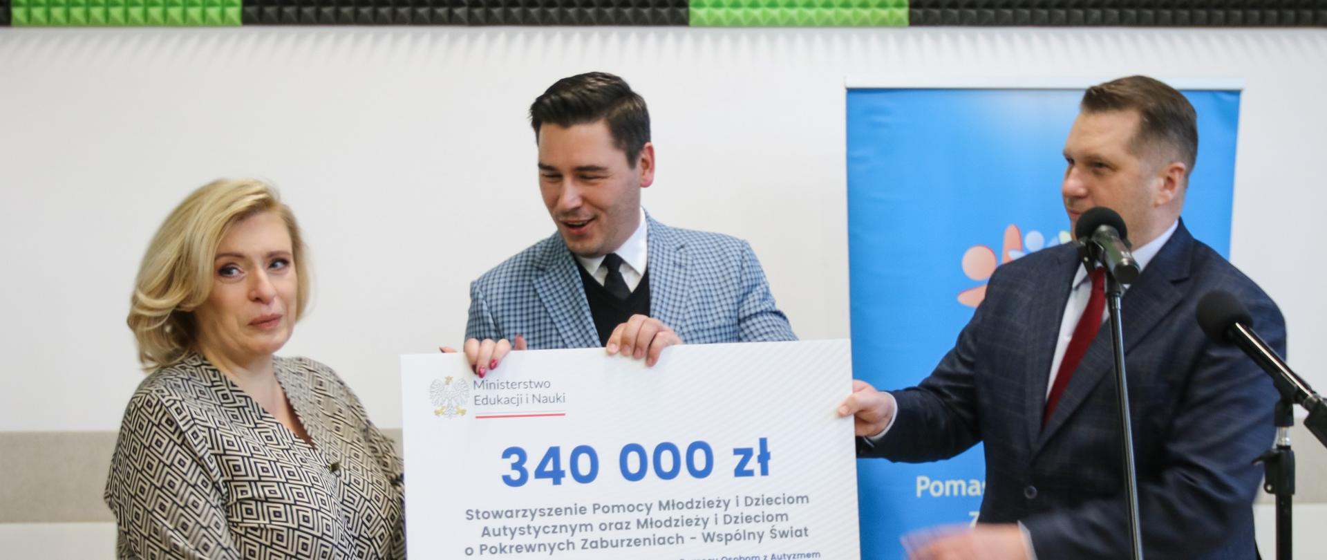Minister, mężczyzna w jasnoszarej marynarce i kobieta w bluzce w szare wzorki stoją i razem trzymają duży symboliczny czek z napisem 340 000 zł, za nimi kolorowy plakat, nad nim ściana w czarno-zielony wzorek.