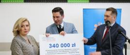 Minister, mężczyzna w jasnoszarej marynarce i kobieta w bluzce w szare wzorki stoją i razem trzymają duży symboliczny czek z napisem 340 000 zł, za nimi kolorowy plakat, nad nim ściana w czarno-zielony wzorek.