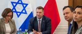 Za stołem siedzą: kobieta w szarej marynarce i białej bluzce, minister Czarnek, mężczyzna w szarym garniturze i niebieskim krawacie i kobieta w bluzce w pionowe biało-czarne paski, za nimi przed białą ścianą flagi Polski i Izraela.