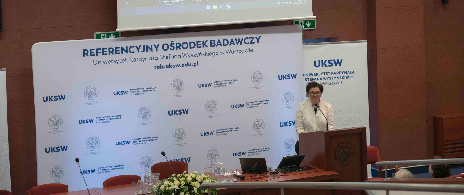 Za drewnianą mównicą stoi kobieta w białej marynarce i mówi do mikrofonu, za nią rozwijany baner z napisem Referencyjny Ośrodek Badawczy, za nim drewniana ściana na której wisi wielki ekran na którym widać ministra Czarnka.