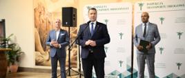 Trzech mężczyzn w garniturach stoi na tle dużego baneru z napisem Inspekcja Transportu Drogowego. Jeden z nich stoi przed mikrofonem i przemawia.