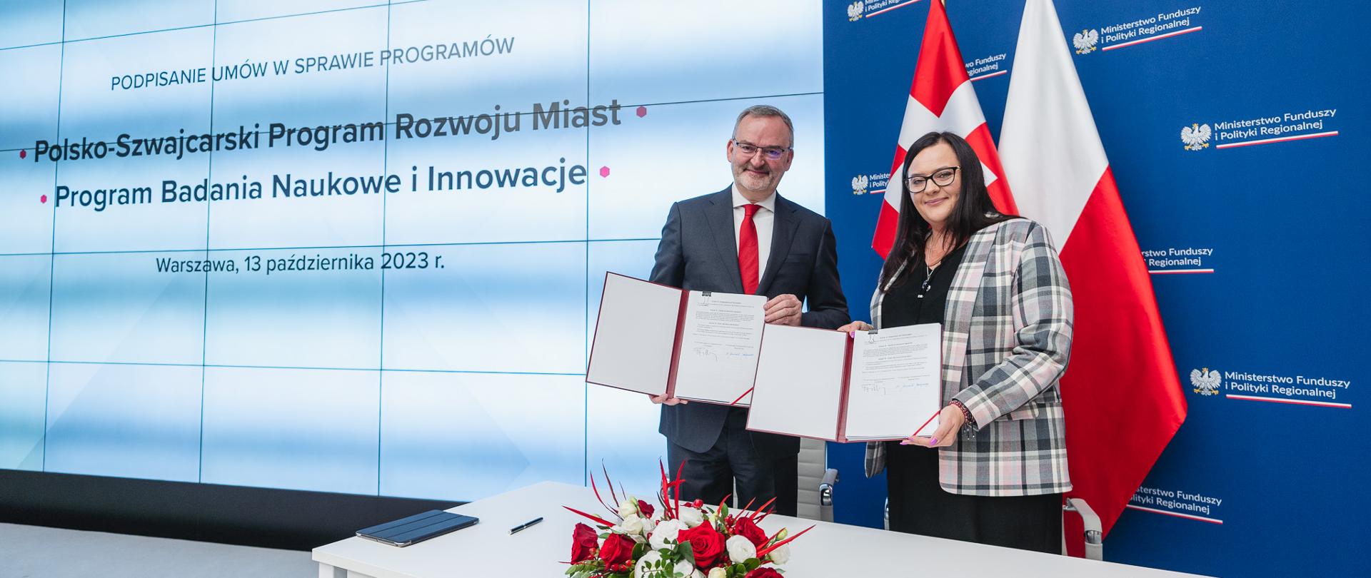 Dwie osoby stoją z umowami w rękach. Pierwsza od prawej stoi uśmiechnięta wiceminister Małgorzata Jarosińska-Jedynak. 