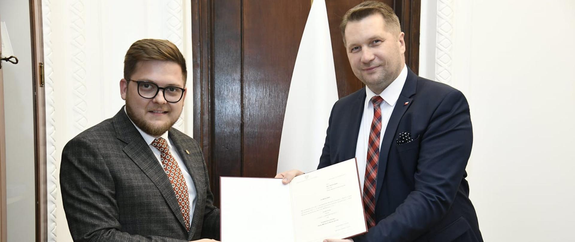 Minister Edukacji i Nauki Przemysław Czarnek wręcza dokument mianowania na przewodniczącego Rady Młodych Naukowców Igorowi Kilanowskiemu. Panowie ubrani są w garnitury. W tle biało-czerwona flaga