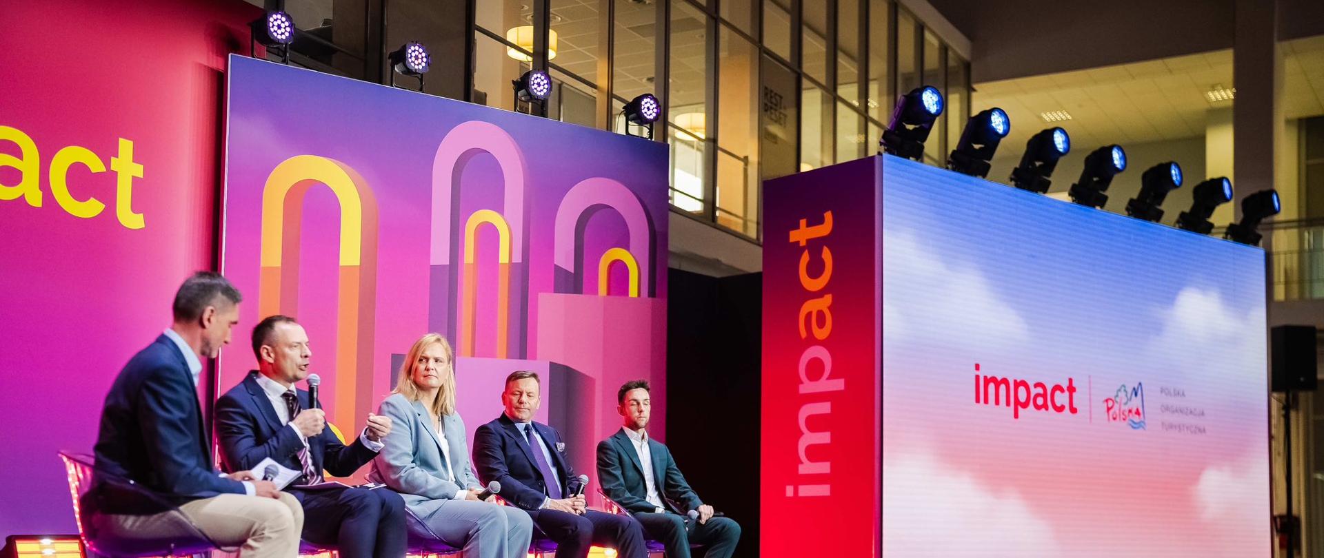 Piotr Borys podczas panelu na forum IMPACT. Po lewej stronie siedzą goście, po prawej stronie duży ekran z logiem POT oraz IMPACT