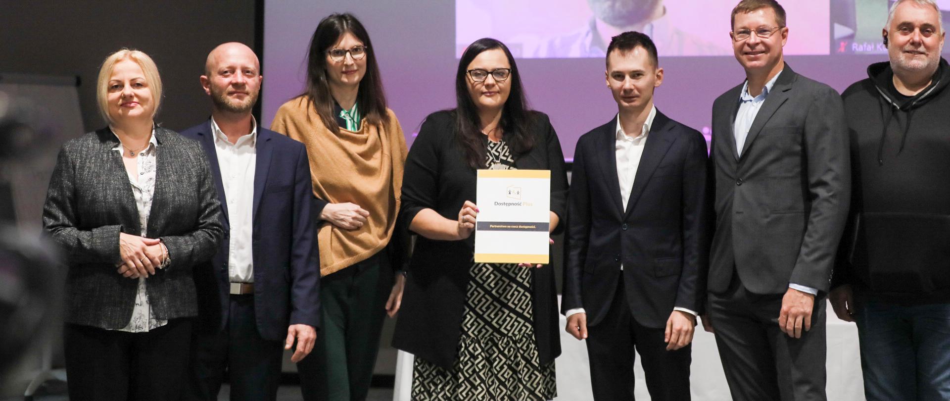 Grupa osób pozuje do zdjęcia. Wśród nich wiceminister Małgorzata Jarosińska-Jedynak z dokumentem w ręku.