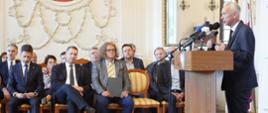 Widok na salę, na krzesłach ustawionych w rzędy siedzą ludzie, w pierwszym rzędzie minister Czarnek obok mężczyzny w jasnoszarym garniturze, z boku mównica za którą stoi mężczyzna w czarnym garniturze i mówi do kilku mikrofonów.