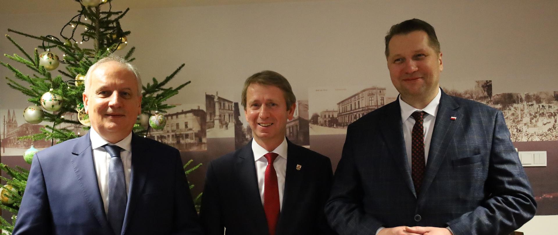 Minister Czarnek i dwóch mężczyzn w garniturach stoją pod ścianą, na której wiszą czarno-białe obrazki z różnymi budynkami, obok choinka z bombkami.