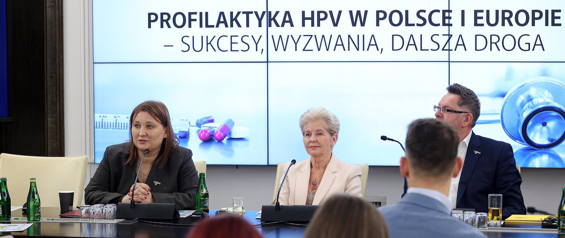 Ujęcie prezydium podczas konferencji poświęconej profilaktyce HPV. Trzy osoby (dwie kobiety i jeden mężczyzna) siedzą za długim, ciemnym stołem. Kobieta pośrodku, ubrana w jasną marynarkę, wydaje się przemawiać. W tle widoczny jest duży ekran projekcyjny z tytułem: "KONFERENCJA PROFILAKTYKA HPV W POLSCE I EUROPIE – SUKCESY, WYZWANIA, DALSZA DROGA". W tle na ekranie widoczne są również elementy graficzne związane z medycyną, takie jak pigułki, strzykawki i stetoskop. Na stołach widać mikrofony, notatniki, butelki wody i materiały konferencyjne.