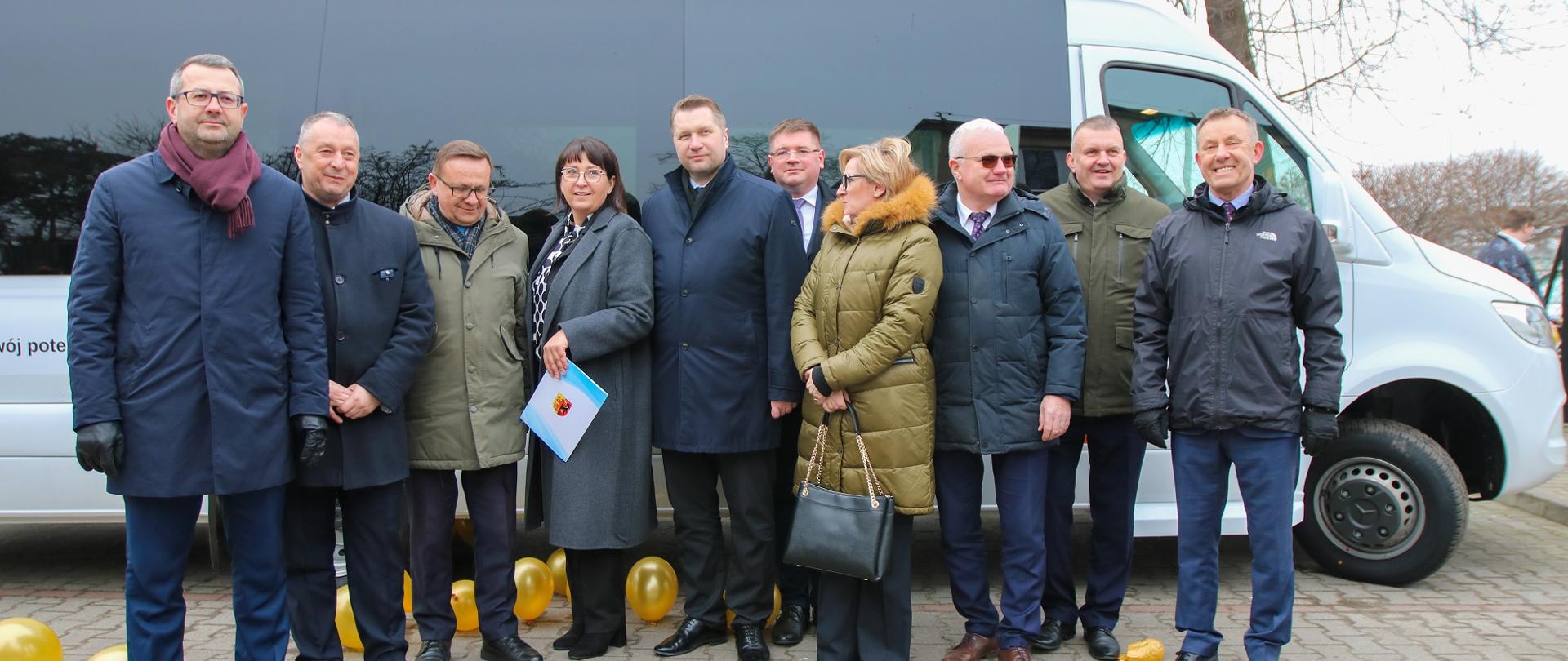 Grupa ludzi z ministrem Czarnkiem pośrodku stoi na wyłożonym szarą kostką podwórku pod białym autokarem.
